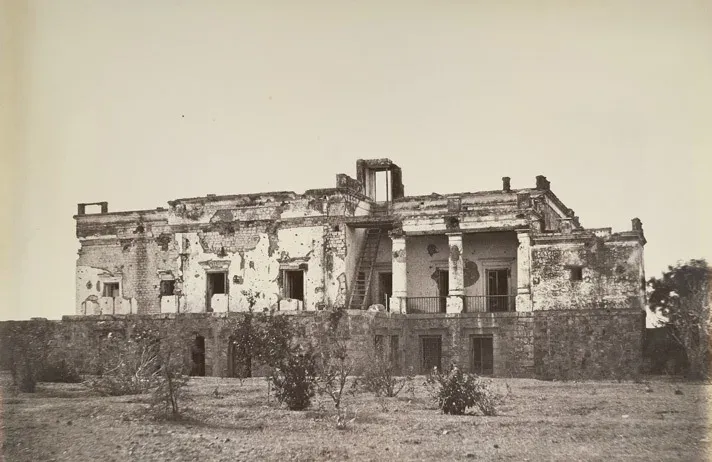 दो मंज़िला पुराना घर, दीवारों पर अनगिनत निशान, ऊपर टूटी छत और एक सीढ़ी; सामने खुला मैदान।