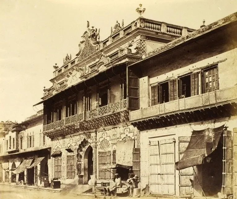 पुरानी दिल्ली की एक सजी हुई हवेली: नक़्क़ाशीदार छज्जे और जालियाँ, नीचे दुकानें और चबूतरे पर बैठे कुछ लोग।
