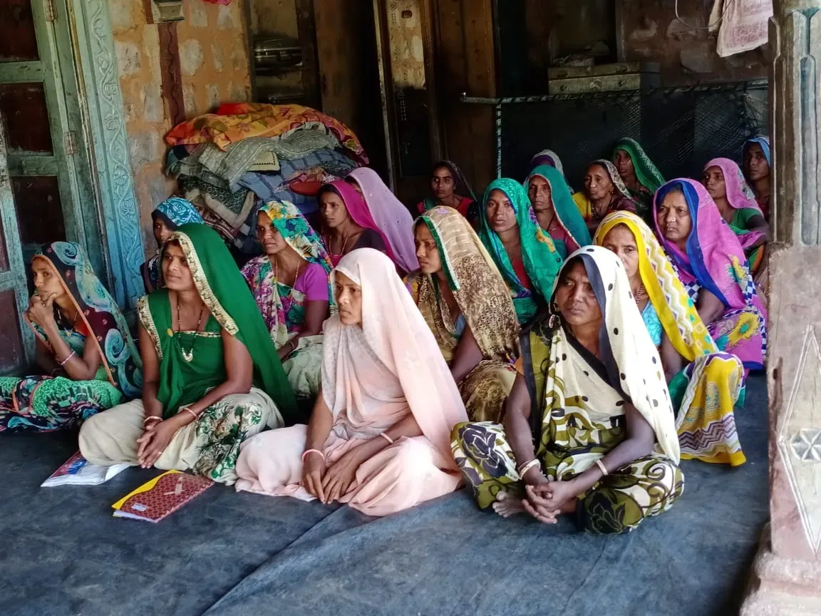 रंग-बिरंगी साड़ियों में कई महिलाएँ एक कमरे में दरी पर बैठी हैं; आगे दरी पर कॉपियाँ रखी हैं।